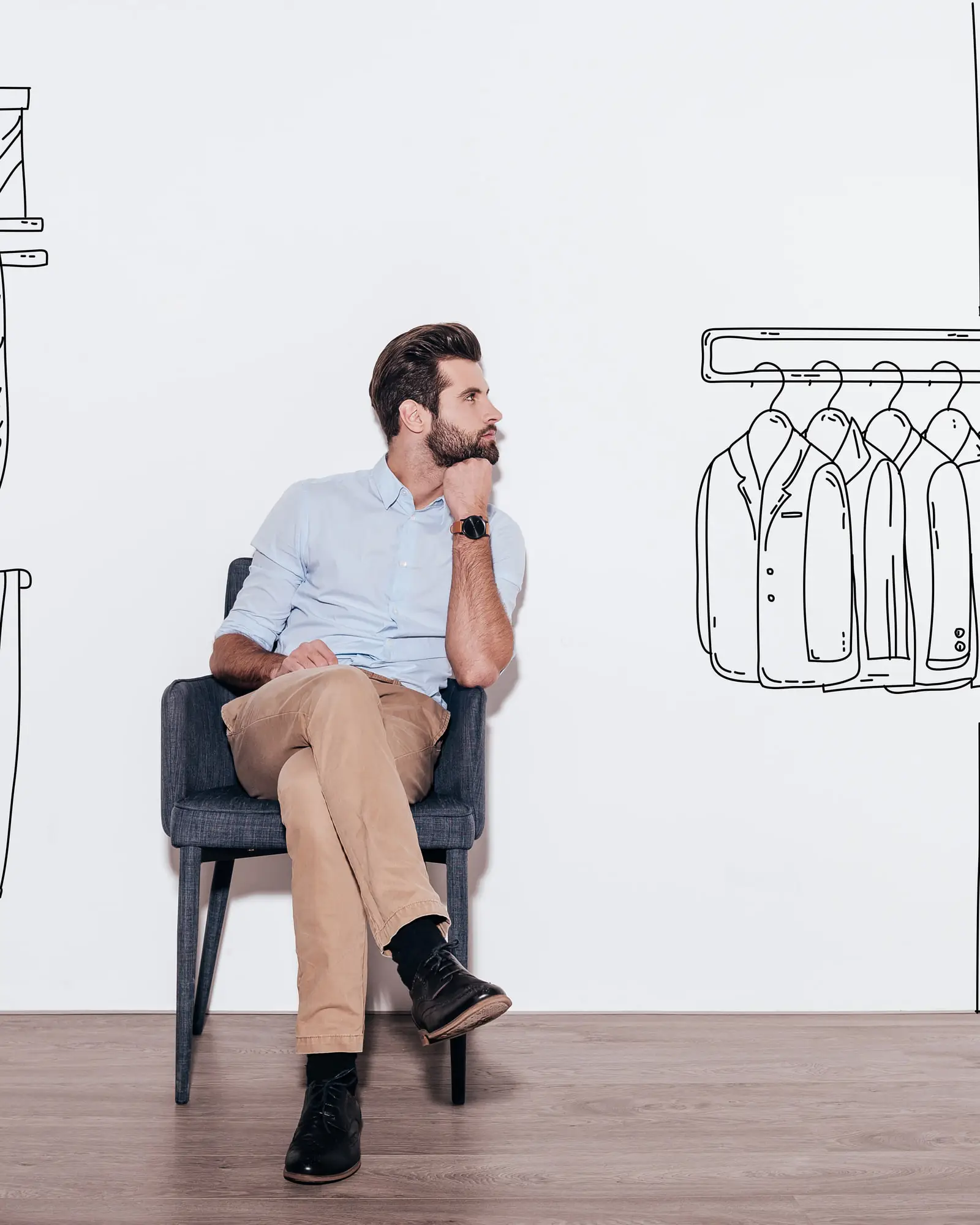 A man sitting in an armchair against a white wall, looking at an illustrated walk-in wardrobe drawn around him