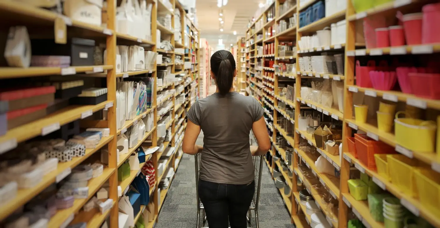 A shopper pushing a cart down a narrow store aisle lined floor to ceiling with storage boxes, baskets and bins